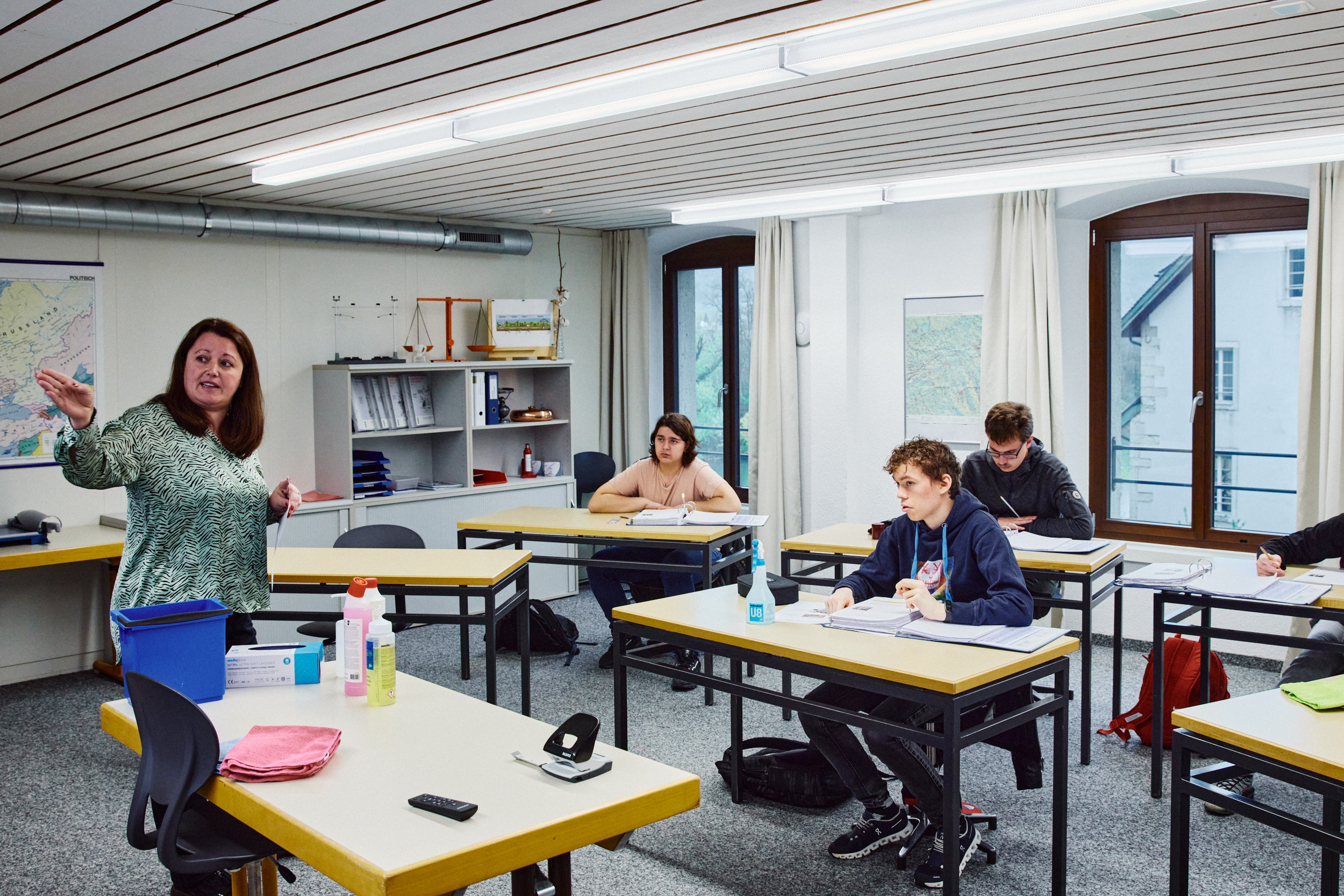 Unterrichtssituation im hellen Schulraum am Standort Aarau, Lehrperson erklärt drei Lernenden etwas im Unterricht